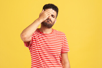 Unhappy forgetful man with beard in striped t-shirt making facepalm gesture keeping hand on head,...
