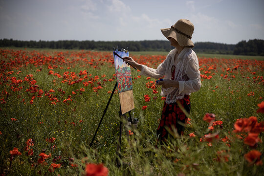 Mature woman artist paints a picture from nature on a field of blooming poppies. Portrait of the artist at work