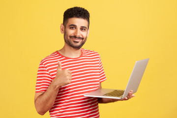 Positive smiling man freelancer with beard in striped t-shirt showing thumbs up standing and holding laptop, satisfied with teleworking, likes his job. Indoor studio shot isolated on yellow background