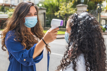 selective focus of a Latina teacher with a mask checking the temperature of a teenage student with an infrared digital thermometer