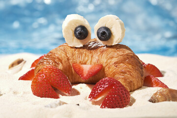 Fun Food for kids. Cute crab croissant with fruit for kids breakfast on the sea sand beach