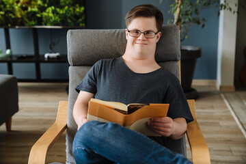 Young man with down syndrome reading book while sitting in armchair