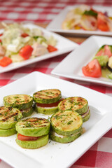 Apetizer from tomatoes, zucchini and mozzarella cheese served on a white plate with other dished on the table.