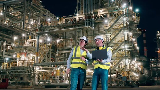 Two engineers with a laptop are talking inside of the oil refinery