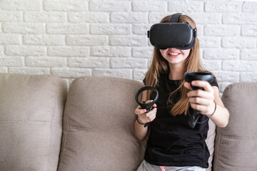 Girl wearing virtual reality VR simulator playing with joysticks at home