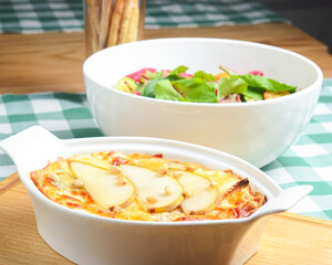Cottage cheese and potato casserole with pears served in a white casserole dish and vegetable salad on the background.