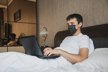A man is sitting in bed in his bedroom and working at a laptop with a medical mask on his face