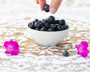 Hand taking a blueberry from a ceramic bowl of fresh blueberries placed on a wooden table covered with  a white lace tablecloth and fresh flowers.