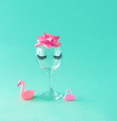 Glass cup with eyelashes, pink bow and women's accessories. Turquoise background. Minimalism.