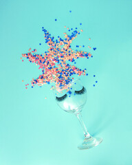 A glass with eyelashes from which he exposes sequins. A party, a women's celebration. Blue background. Minimal composition.