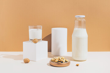 Glass and a bottle with vegan milk from various nuts with white geometric figures on sunlit background. Dairy and lactose free substitute drink. Healthy food ingredient concept. Trendy colors.