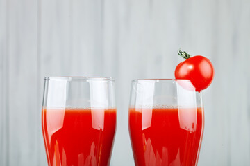 Two glasses with tomato juice and a small tomato on a bright background.