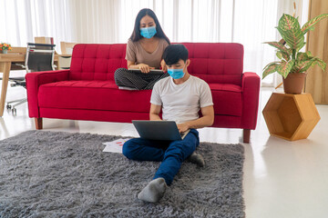 Obraz premium Young couple wearing face masks protection covid 19 use laptop working from home while sitting on sofa during quarantine coronavirus pandemic new normal policy implemented. Lifestyle outbreak concept
