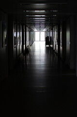 Dark empty long indoor building corridor leading to glass door in the distance. Daylight outside, no people
