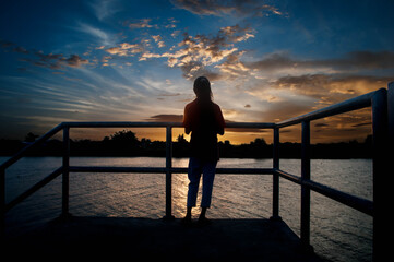 Fototapeta premium A little girl stands and watches the sun go down to the beautiful riverside sky.