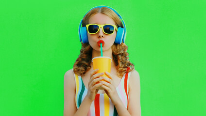Summer portrait of stylish young woman listening to music in headphones and drink fresh juice on green background