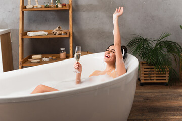 happy woman in headphones listening music and holding glass of champagne while taking bath