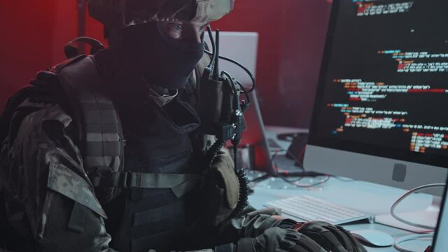 Handheld slowmo tracking shot of masked special forces officer in uniform sitting at desk in hackers hideout and looking through data on computer during raid