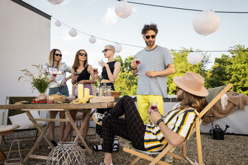 Young group of stylish people having a festive dinner on the roof terrace. Hipsters hanging out and having great summertime together outdoors