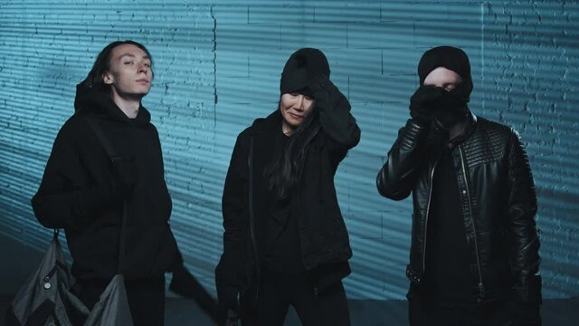 Portrait dolly-in shot of three young female and male robbers taking off their balaclavas and looking at camera