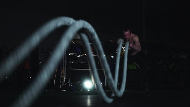 Muscular man with naked torso exercising with battle ropes in dark gym, equipment on background. Athlete doing high intensity interval training. Concept of sport