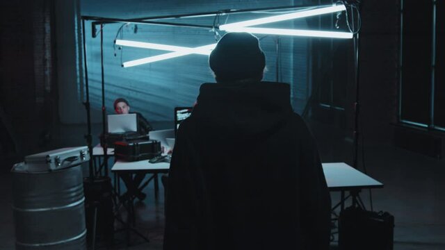 Slowmo tracking of young man in hoodie and beanie hat walking in dark industrial space turned into hackerspace He is sitting down at his desk typing on computer