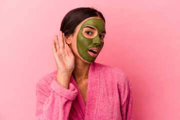 Young caucasian woman wearing a bathrobe and facial mask isolated on pink background trying to listening a gossip.
