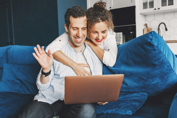 Happy millennial man use laptop at home on sofa in living room waving hello to friends and family remotely during quarantine. Joyful smiling woman wife standing by hugging husband with warmth and love