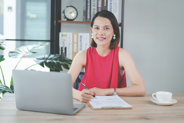 Obraz premium A cheerful middle-aged Asian businesswoman in relax casual dress working at home, checking email on computer laptop, writing on financial accounting document paper. Stock photo