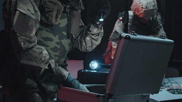Slowmo tilt up shot of special forces officers in uniforms raiding hackers hideout One of them is checking laptop while other one is looking into briefcase