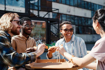 Diverse group of contemporary young people enjoying pizza outdoors, scene lit by sunlight, copy space
