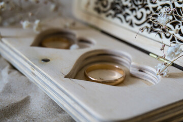 Photo of a wooden box with a pair of silver wedding rings on it. A heart is carved in the box as a symbol of eternal love. The ring box is surrounded by white delicate flowers, the composition is set 