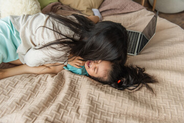 Mother kissing cheerful asian daughter near blurred laptop on bed
