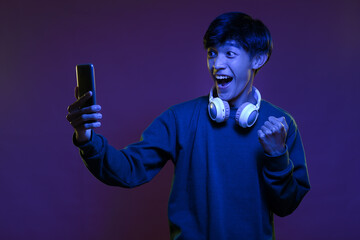 Portrait of excited handsome young asian man looking at the mobile phone wearing headphone in neon...