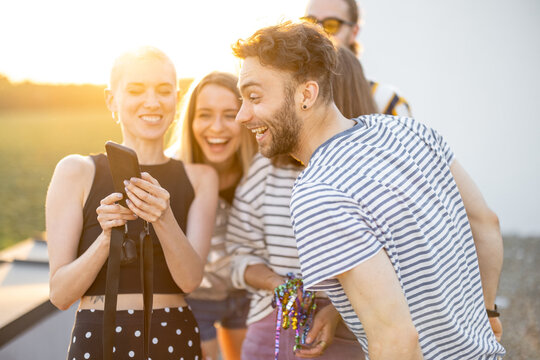 Young stylish friends have fun, showing something on phone and laugh outdoors. Hipsters, millennials hang out at rooftop terrace on sunset