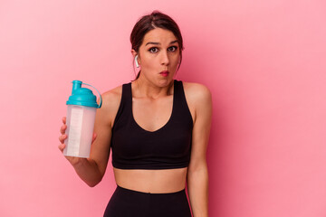 Young caucasian woman drinking a protein shake isolated on pink background shrugs shoulders and open eyes confused.
