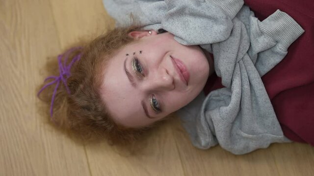 Top view of joyful redhead Caucasian woman with grey eyes looking at camera smiling lying on floor at home. Portrait of relaxed cheerful millennial hipster posing indoors. Lifestyle concept