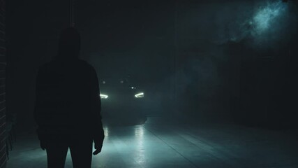 Slowmo tracking shot of silhouette of unrecognizable criminal standing in dark industrial space while car with two masked terrorists driving towards him One of them is exiting car