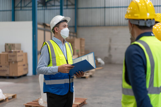 Asian Male Supervisor Engineer In Wearing Helmet Safety And Hygienic Mask To Protect Coronavirus Stands With Documents Explaining Action Plan To Teamwork At Warehouse Factory Industrial.