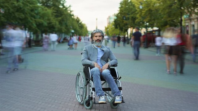 Time lapse portrait of disabled mature man sitting in wheelchair in busy pedestrian street while people are passing by. Disability and life concept.