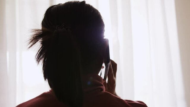 Asian woman talking on cell phone near window with sunlight.