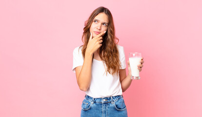 young pretty woman thinking, feeling doubtful and confused and holding a glass of milk