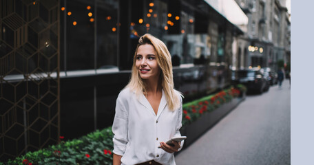 Fototapeta premium Smiling woman browsing smartphone near modern buildings on street