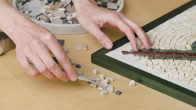 Male hands create mosaics pattern using tweezers and small pieces of granite stone. Master making handicraft in creative workshop. Interesting hobby, art, artwork. Close up. Slow motion ready 59.94fps