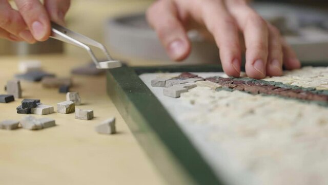 Male hands create mosaics pattern using tweezers and small pieces of granite stone. Master making handicraft in creative workshop. Interesting hobby, art, artwork. Close up. Slow motion ready 59.94fps