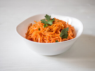 Spicy Asian salad with grated carrots and parsley. Korean carrots in a white bowl on a white backgroung. Vegetarian diet. Healthy food concept. View from above. Horizontal.