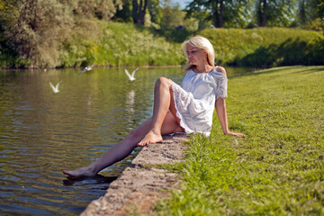 Naklejka na ściany i meble Pretty dreaming young woman sitting near lake in summer park in a warm sunny day. blonde woman sitting near pond
