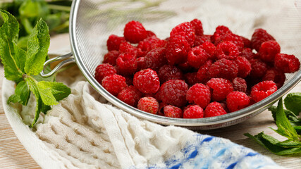 A lot of ripe raspberries in a colander after washing, lies and dries on a wooden table with a mint and a tea towel