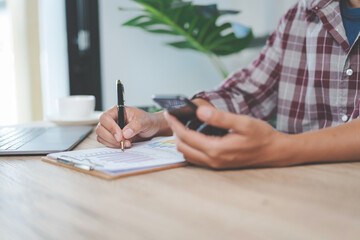 Close-up businessman holding a pen, smartphone and pointing at financial graph checking business report on wooden desk with computer laptop besided at home. Stock photo