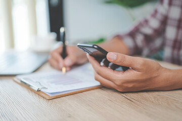Fototapeta premium Close-up businessman holding a pen, smartphone and pointing at financial graph checking business report on wooden desk with computer laptop besided at home. Stock photo